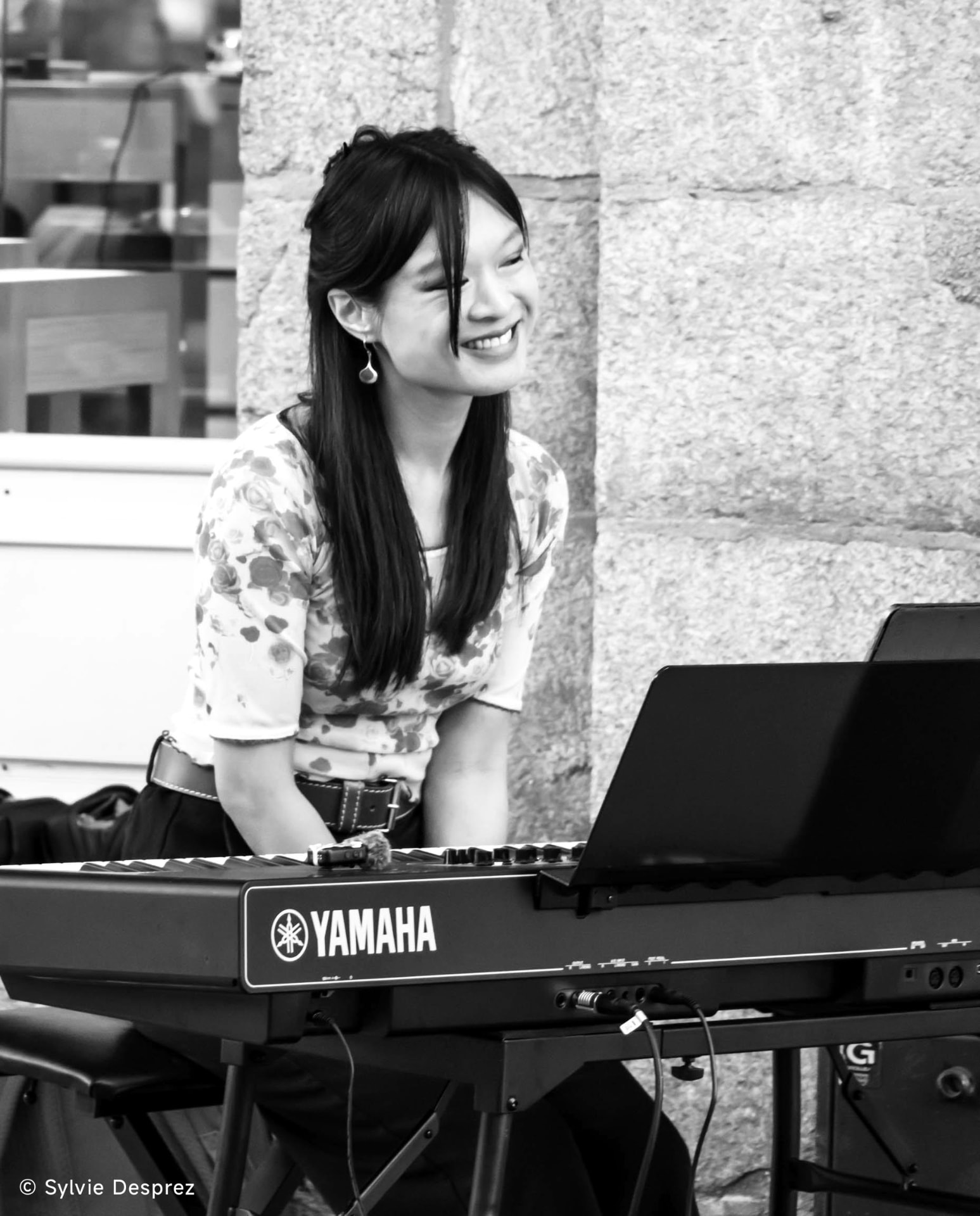 En noir et blanc, Héléna Riols rit, penchée sur son clavier Yamaha devant un mur de pierre.