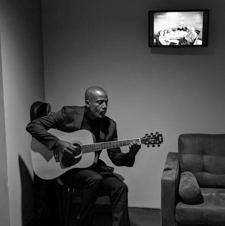 Jean-Marc assis avec sa guitare dans une salle de cérémonie, en noir et blanc.