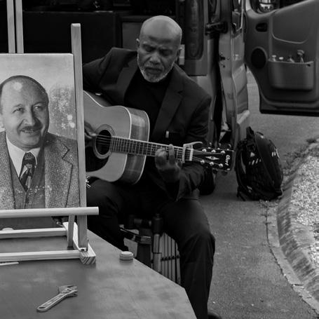 Jean-Marc joue de la guitare lors d’un hommage funéraire, près d’un portrait, en noir et blanc.