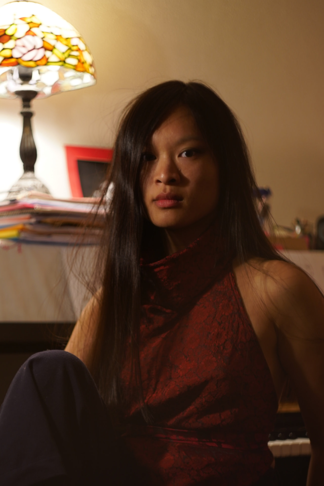 Portrait d'Héléna Riols en haut rouge, longs cheveux noirs, près d'une lampe à abat-jour en vitrail.